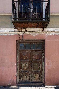 Küçük dövülmüş demir bir balkonun altında eski, kahverengi ahşap bir kapının dikey görüntüsü solmuş pembe alçı bir duvara karşı, kentsel bozulmayı ve tarihi dokuları vurguluyor..