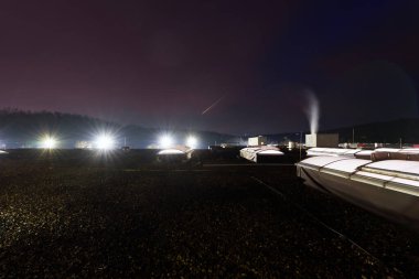 Yıldızlı gökyüzüne karşı sessiz fabrikalar, fabrika siluetleri ve yumuşak şehir parıltılarının yer aldığı gece manzarası, gökyüzü altındaki endüstriyel çatılar, uzak şehir aydınlatması ve gölgelerinden hafif aydınlatmalardır.
