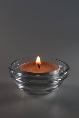Tarafsız gri bir arkaplanda şeffaf bir cam kasede parlayan bir ışık mumu. Görüntü, barış, maneviyat, meditasyon veya hatırlama temaları için mükemmel, parlak bir alev ve yumuşak bir aydınlatma içerir. Metin için kopyalama alanı içerir.