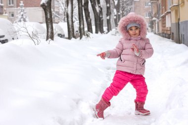 Pembe kışlık elbiseli küçük bir kız karla kaplı bir şehir sokağında yüzünde neşeli duygularla mutlu bir çocukluk ve aktif bir kış yaşam tarzıyla duruyor.