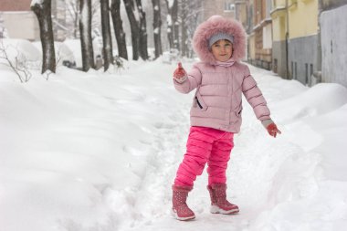 Pembe kışlık elbiseli küçük bir kız karla kaplı bir şehir sokağında yüzünde neşeli duygularla mutlu bir çocukluk ve aktif bir kış yaşam tarzıyla duruyor.