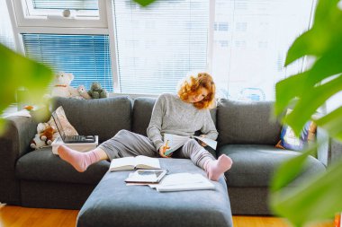 Kıvırcık saçlı genç bir kadın not defterine not alıyor, evde e-öğreniyor, online öğrenci.