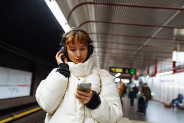 Yeraltı toplu taşımacılığındaki kışlık ceketli genç kadın cep telefonu kullanıyor
