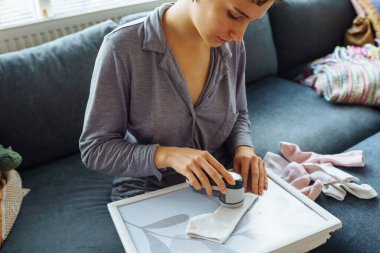 Kadın evde kıyafetlerle ilgileniyor, aletle ipliği kesiyor, eşyaları sürekli kullanıyor, çevre dostu.