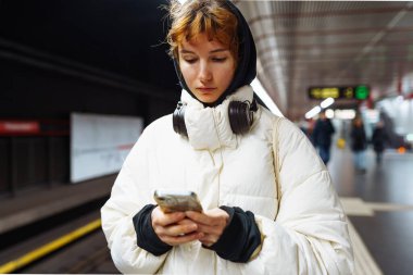 Yeraltı toplu taşımacılığındaki kışlık ceketli genç kadın cep telefonu kullanıyor