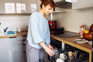 Bulaşık makinesine kirli bulaşık koyan genç bir kadın. Mutfakta ev işi.
