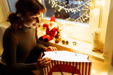 Genç kadın Noel hediyeleriyle dolu çantaların yanında oturuyor, arka planda çelenkler parlıyor, tatil beklentisi içinde.