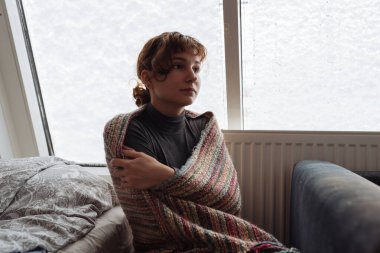 Genç bir kadın pencerenin yanında, battaniyeye sarılmış bir şekilde yatağında oturuyor. Radyatörün yanında ısınıyor, merkezi ısıtma çalışmıyor.