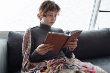 Genç kadın yumuşak kanepeye oturur, sıcak bir battaniyeye sarınır, karla kaplı pencerenin yanında, kitap okur, kitap ve defterleri elinde tutar.