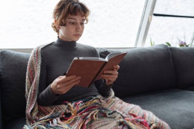 Genç kadın yumuşak kanepeye oturur, sıcak bir battaniyeye sarınır, karla kaplı pencerenin yanında, kitap okur, kitap ve defterleri elinde tutar.