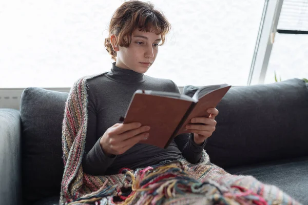 Genç kadın yumuşak kanepeye oturur, sıcak bir battaniyeye sarınır, karla kaplı pencerenin yanında, kitap okur, kitap ve defterleri elinde tutar.