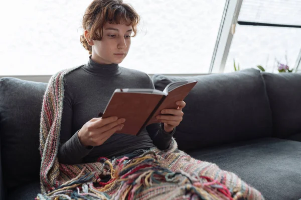 Genç kadın yumuşak kanepeye oturur, sıcak bir battaniyeye sarınır, karla kaplı pencerenin yanında, kitap okur, kitap ve defterleri elinde tutar.