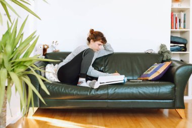 Genç kadın rahat deri koltukta oturur, not defterine not alır, kendi kendine çalışır, evde uzaktan öğrenir.