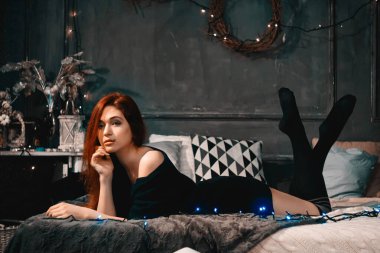 Kızıl saçlı kadın yeni yılı kutlamak için rahat bir evde yalnız kalır ve Noel ışıkları ve çelenklerle çevrili gri bir yatakta yatar.. 
