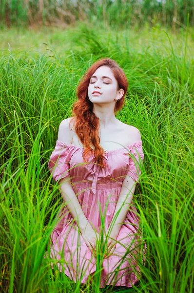 Outdoor portrait of young redhead woman in field of green grass with pink peony. Woman\'s Day. Female spring, summer fashion concept.