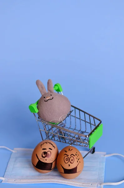 Creative Easter eggs with Corona virus (COVID19) protection concepts. Diverse chicken eggs on mini shopping cart with doodle faces wearing medical masks on blue background. Flat lay, top view.