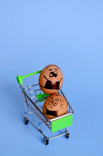 Creative Easter eggs with Corona virus (COVID19) protection concepts. Diverse chicken eggs on mini shopping cart with doodle faces wearing medical masks on blue background. Flat lay, top view.