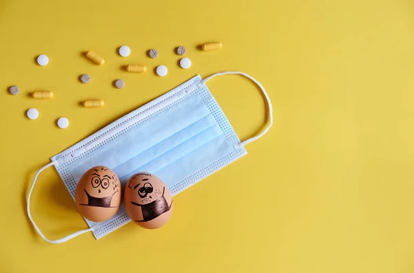 Creative Easter eggs with Corona virus (COVID19) protection concepts. Chicken eggs with doodle faces wearing medical masks with pills on yellow background. DIY. Flat lay, top view, copy space.