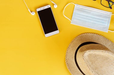 Covid-19 ve yeni normal kavramlardan sonra seyahat et. Tıbbi yüz maskesi, el jeli dezenfektan, kulaklık ve sarı arka planda plaj şapkası. Koronavirüsü önlemek için yaratıcı fikirler.