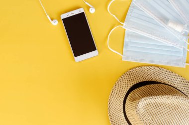 Covid-19 ve yeni normal kavramlardan sonra seyahat et. Tıbbi yüz maskesi, el jeli dezenfektan, kulaklık ve sarı arka planda plaj şapkası. Koronavirüsü önlemek için yaratıcı fikirler.