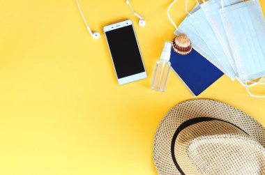 Covid-19 ve yeni normal kavramlardan sonra seyahat et. Tıbbi yüz maskesi, el jeli dezenfektan, kulaklık ve sarı arka planda plaj şapkası. Koronavirüsü önlemek için yaratıcı fikirler.