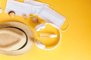 Covid-19 ve yeni normal kavramlardan sonra seyahat et. Tıbbi yüz maskesi, el jeli dezenfektan, kulaklık ve sarı arka planda plaj şapkası. Koronavirüsü önlemek için yaratıcı fikirler.