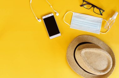 Covid-19 ve yeni normal kavramlardan sonra seyahat et. Tıbbi yüz maskesi, el jeli dezenfektan, kulaklık ve sarı arka planda plaj şapkası. Koronavirüsü önlemek için yaratıcı fikirler.