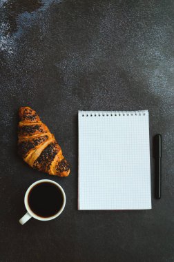 İş adamı sabah not defteri, bir fincan sıcak kahve ve siyah arka planda tatlı kruvasanla çalışıyor. Üst görünüm, kopyalama alanı, modelleme. Düz yatıyordu. Modern kahvaltı yemekleri..