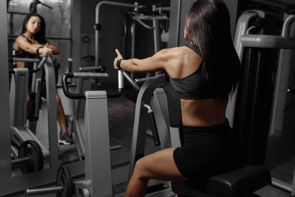 Vista posterior de la fuerte mujer deportiva muscular que se ejercita en la máquina de ejercicio delante del espejo —  Fotos de Stock