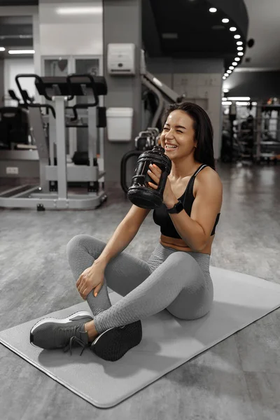 Gülümseyen çekici sportif kız halının üzerinde oturuyor, mola veriyor, duraklıyor, spor salonunda su içiyor.