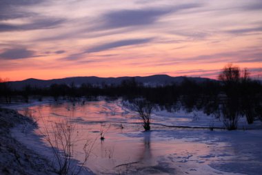 Buz gibi nehir yüzeyi gün batımı manzarası. Kar ve çalılar nehir kıyısında siluetler ve ufuktaki dağlar alacakaranlık şafağına karşı canlı pembe bulutlu gökyüzü