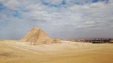 Giza Piramitlerinin çarpıcı manzarasını keşfedin. Tarihi Mısır piramitlerini, bulutlarla dolu mavi bir gökyüzünün arka planına bakarak, antik harikaların özünü yakalayın..