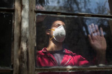 Üzgün, endişeli, N95 koruyucu tıbbi maske takan genç adam pencereden bakıyor, Coronavirus COVID-19 salgını, kendini izole etme ve karantina, sosyal fiziksel mesafe, evde güvende kal, umut ve inanç