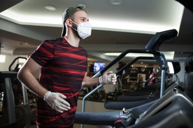 Spor salonunda koşu bandında koşan, koruyucu maske takan ve lastik eldiven takan genç bir adam, COVID-19 salgın hastalığın yayılmasının önlenmesi ve Coronavirus 'un ABD' de kilit altında tutulma kavramında kalması