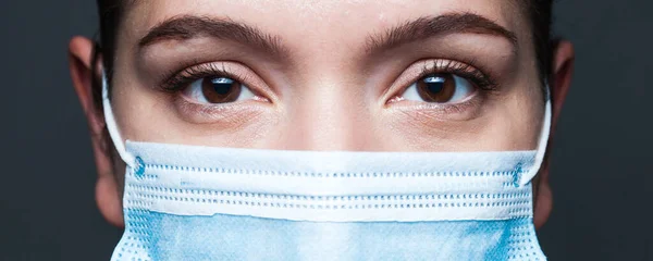 Young caucasian woman wearing blue surgical face mask,closeup widescreen portrait,uncertainty in eyes,isolated on dark gray background,Coronavirus COVID-19 pandemic crisis prevention and protection