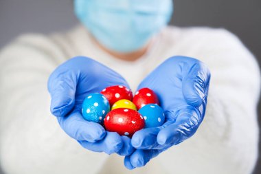 Yüz maskesi takan ve elinde çikolatalı Paskalya yumurtası tutan mavi koruyucu eldivenli kişi, küresel Coronavirus COVID-19 Corona virüsü salgını nedeniyle bayram kutlamaları iptal edildi.