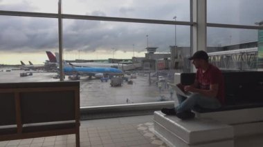 Bir adam havaalanının giden yolcu salonundaki dizüstü bilgisayara bir şey yazıyor. Arka planda uçaklar var. Schiphol Airport, Amsterdam