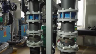 Birkaç siyah yalıtılmış boru, yeşil pompalar ve endüstriyel ekipmanların bulunduğu teknik bir odada gümüş cıvatalı birkaç gri metal flansa bağlıdır..