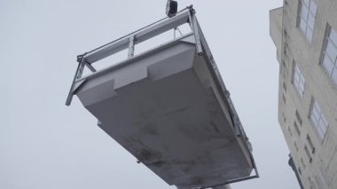 İnşaat veya bakım sırasında gri gökyüzüne karşı vinç kabloları ile asılı büyük endüstriyel metal platformu.