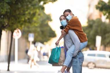 Coronavirus konsepti sırasında alışveriş. Birkaç alışveriş çantası ve tıbbi maske takmış, yaya caddesinde bir kızı sırtında taşıyan yakışıklı bir adam..
