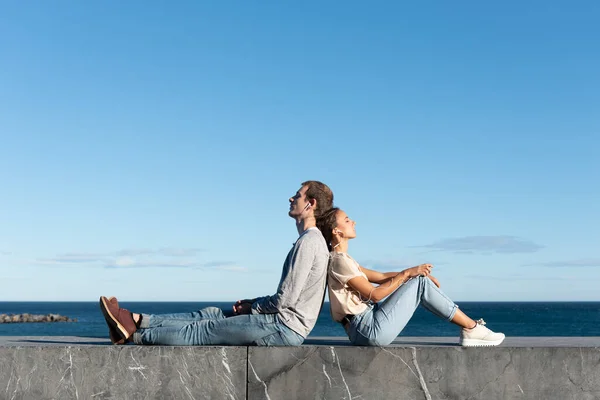 一对穿着休闲的年轻夫妇在阳光明媚的海边用耳机听音乐 背靠背地坐着 — 图库照片