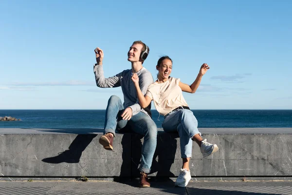 在阳光明媚的日子里 一对年轻夫妇坐在海边的石座上 一边听音乐 一边跳舞 — 图库照片
