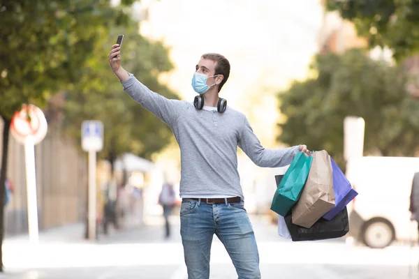 stock image Shopping during Coronavirus concept. A young man wearing a face mask taking a selfie while holding his shopping bags.