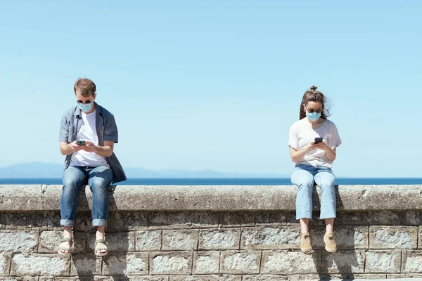マスクをした若者と女性は夏の晴れた日にスマートフォンをチェックします コロナウイルスのパンデミックの概念における社会的距離を保つ — ストック写真