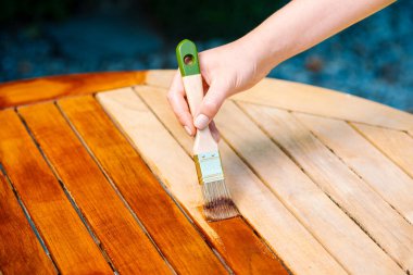 Ahşap bir bahçe masasına cila sürerek ahşabı yağlı boya ile boyamak ve onunla ilgilenmek için bir el fırçası tutuyor.