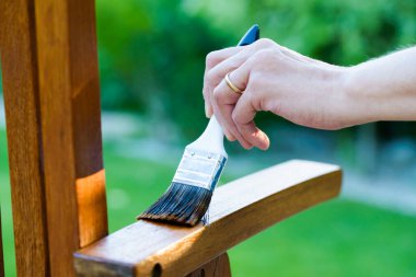 woman hand holding a brush applying varnish paint on a wooden garden chair - painting and caring for wood with oil