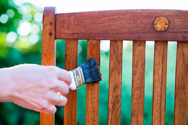 woman hand holding a brush applying varnish paint on a wooden garden chair - painting and caring for wood with oil