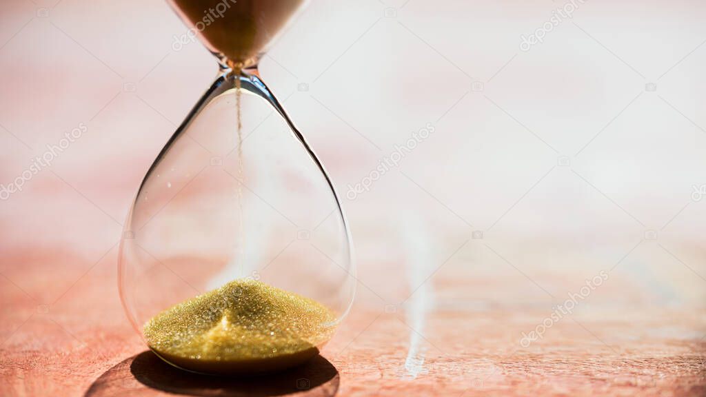 Gold Sand running through the bulbs of an hourglass measuring the passing time in a countdown to a deadline, on a light pastel reddish background with copy space - shallow depth of field