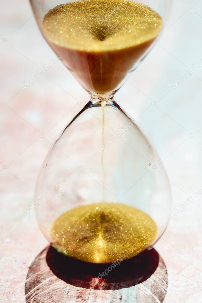 Gold Sand running through the bulbs of an hourglass measuring the passing time in a countdown to a deadline, on a light pastel reddish background - shallow depth of field