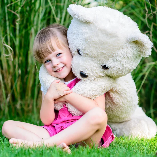 Little cute girl sitting in the grass with large teddy bear